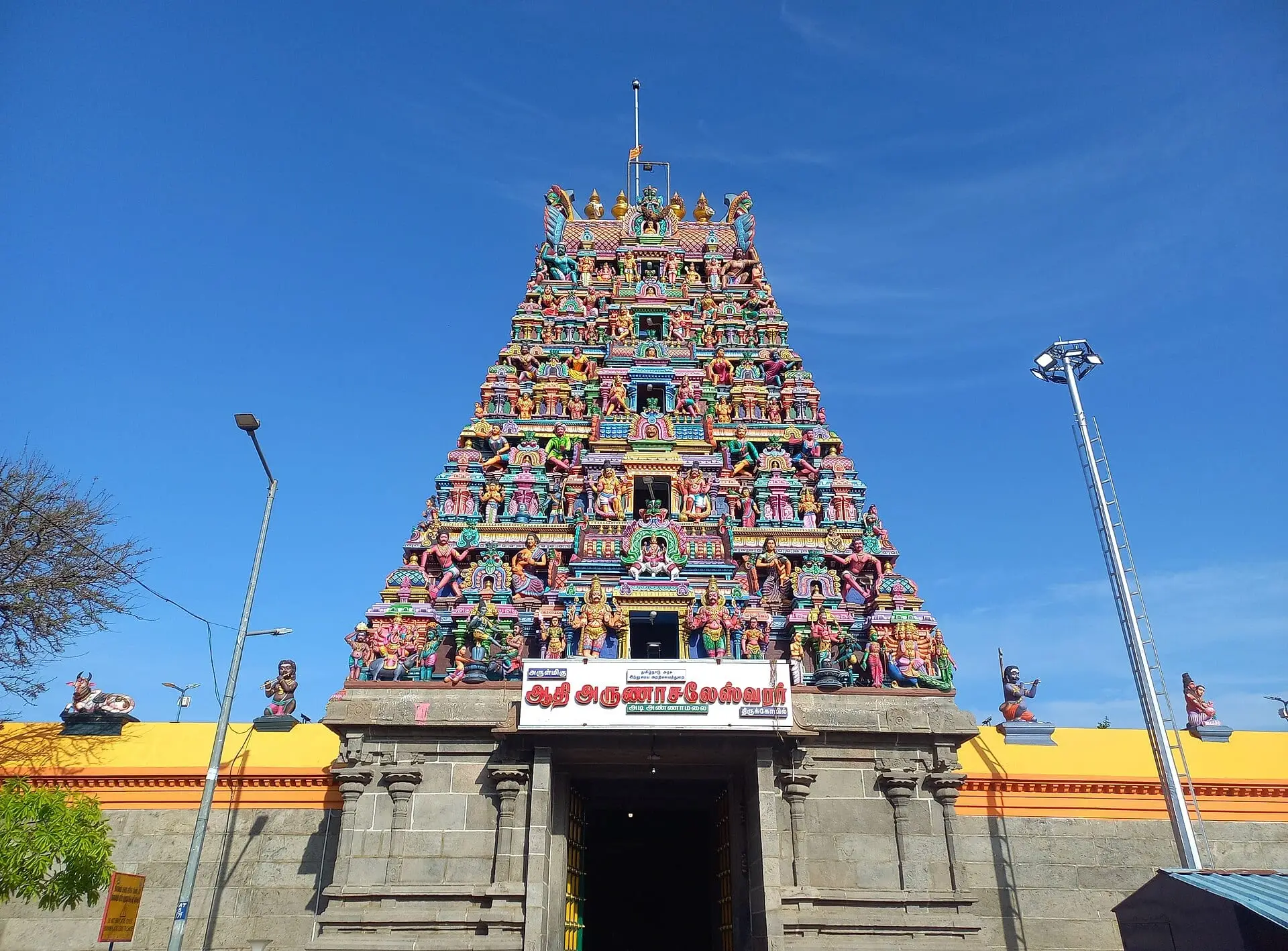 The painted gopuram of Adi Annamalai Temple against a blue sky, with a sign reading Adi Arunachaleswarar